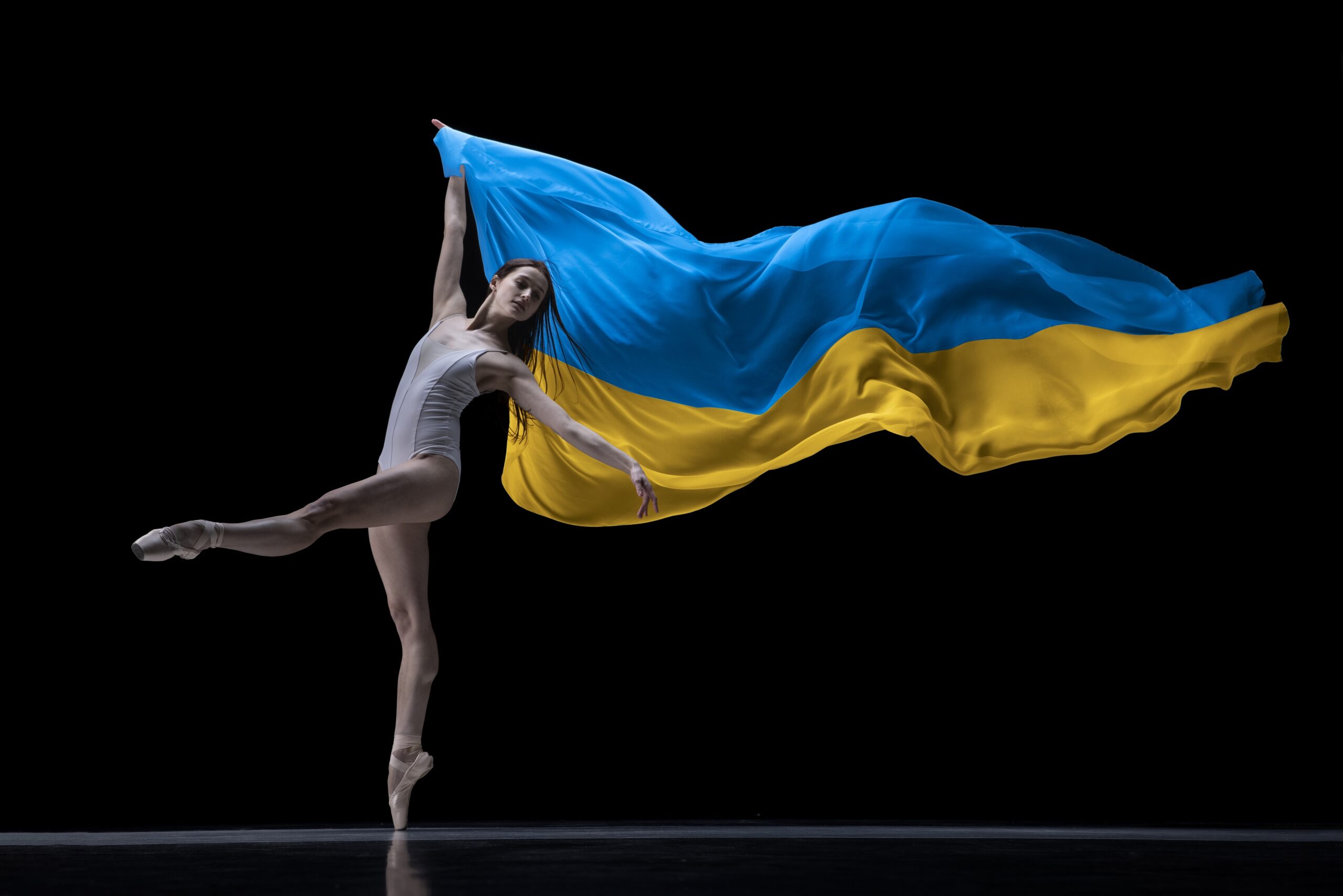 Young girl, ballerina dancing with cloth painted in blue and yellow colors of Ukraine flag on dark studio background.